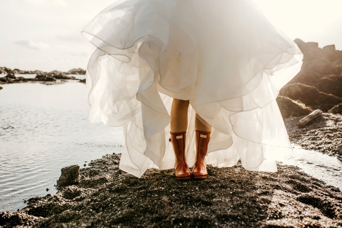 Brides shows off yellow rubber boots while standing along ocean in bridal gown by Shades of White