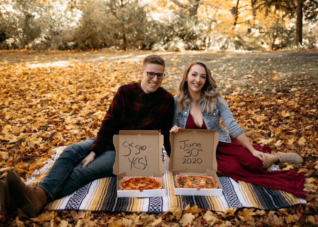 Engaged Couple sit in fall leaves with pizza boxes announcing date