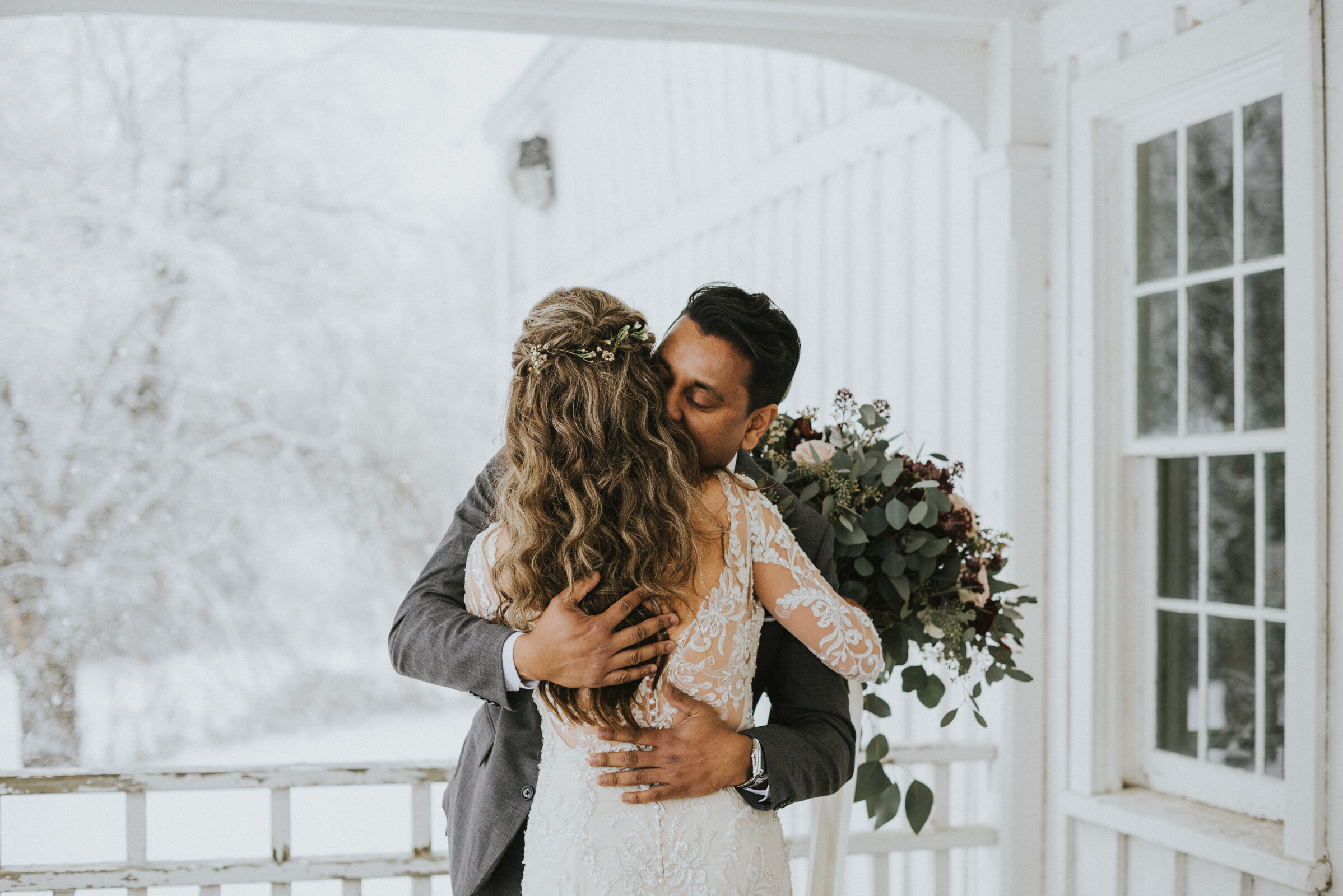 Groom kisses bride during first look on farmhouse porch with snow