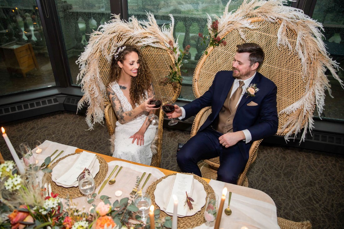Newlyweds sit in large wicker chairs edged in pampas grass in front of sweetheart table by Wild Boheme Decor