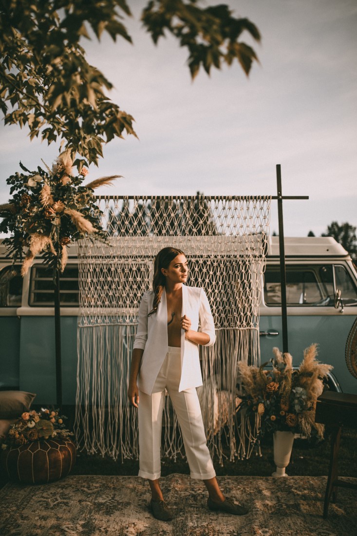 Bride in white Azaria pantsuit in front of macrame backrop and blue VW van