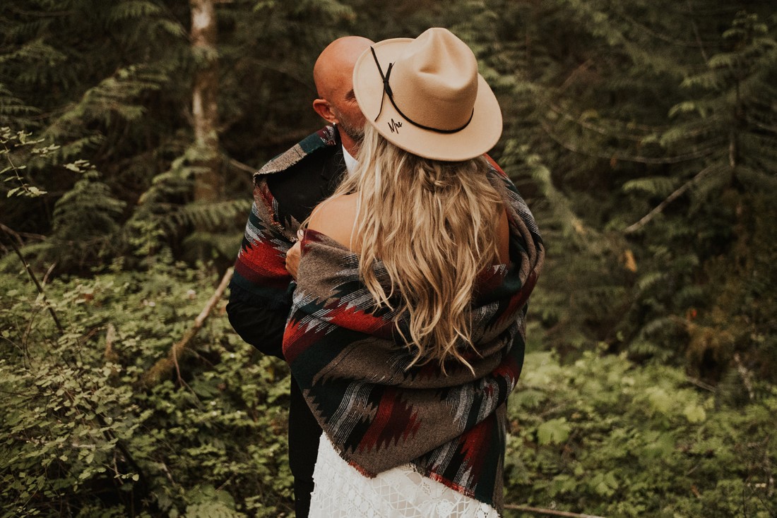 Bride wearing hat and striped blanket kisses her groom in the forest 