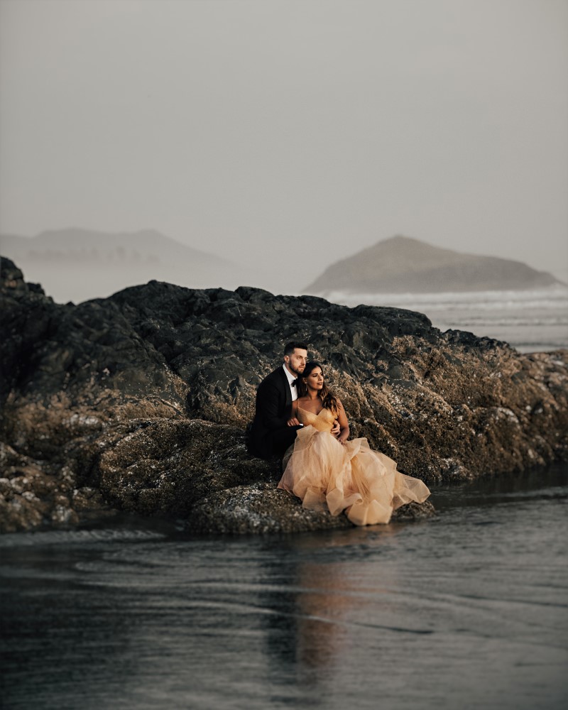 Couple sit on the black rock of Tofino beach in wedding gown and tux