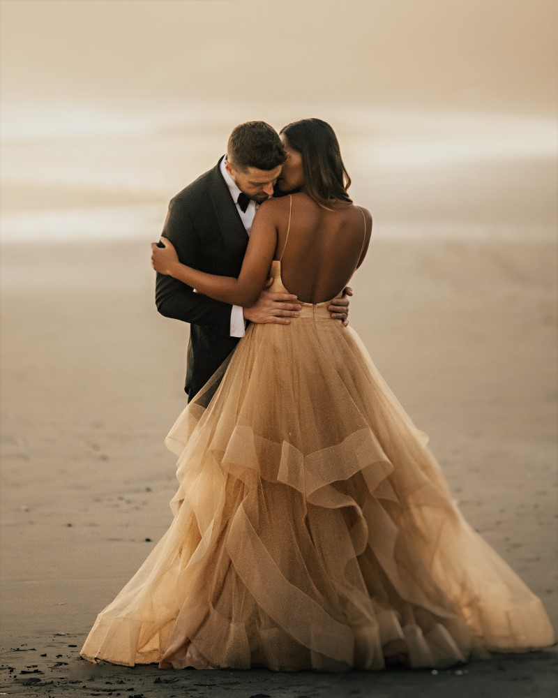 Groom in black tux leans over bride wearing BHLDN peach gown on beach