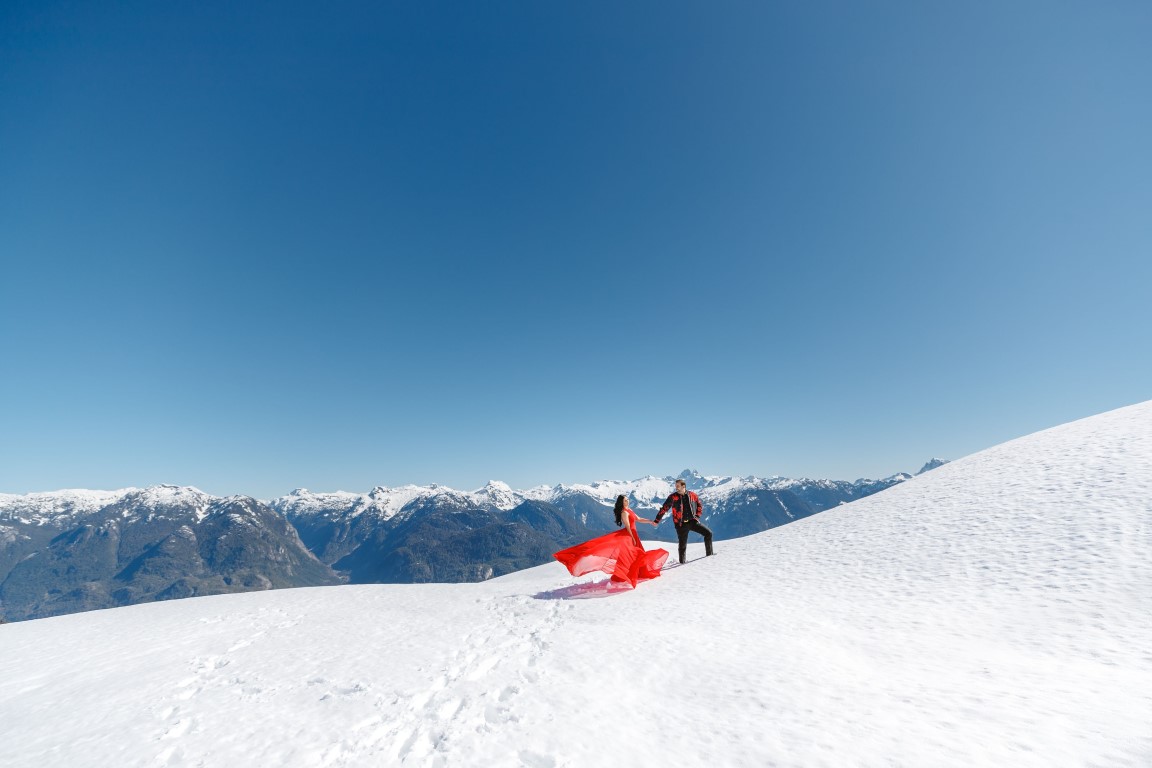 Couple in red gown walk along the top of BC mountains by SoWedding