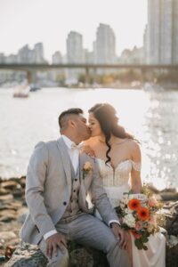 groom and bride sit kissing on large rock at the oceanside